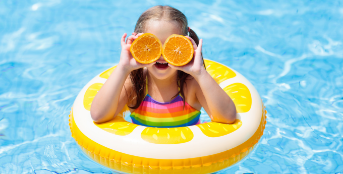 Young girl in a yellow ring in the swimming pool holding two half oranges to her eyes.