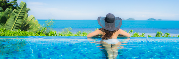 A woman is enjoying a relaxing swim in a beautiful, sunlit pool with a view of the ocean in the background. She is resting on the edge of the pool with her arms and legs in the water, appearing to be at peace and comfortable.